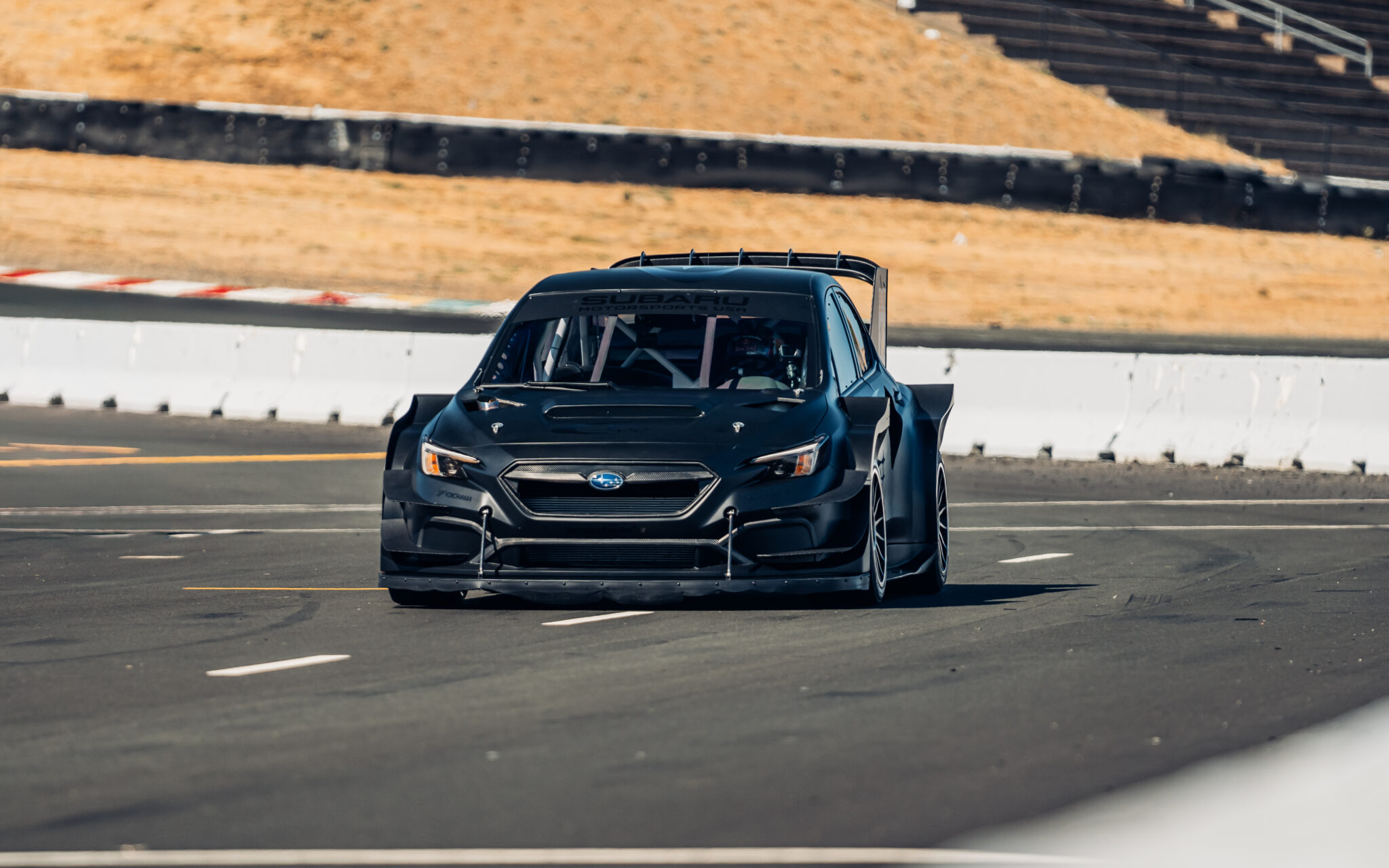 Mobil balap hitam dengan spoiler besar merobohkan arena pacuan kuda di Sonoma Raceway. Langit yang cerah dan rumput kering membingkai pemandangan, menangkap sensasi festival Motorsports Invitational Velocity.