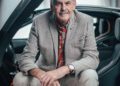 An elderly man with grey hair and a mustache sits in a modern car, reflecting the pinnacle of automotive design, with open doors. He's wearing a light grey blazer, floral shirt, and beige pants inside a well-lit modern building reminiscent of Goodwood's elegance.