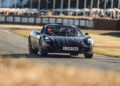 A sleek black sports car, reminiscent of Gordon Murray's iconic automotive designs, speeds around the track at Goodwood. With two occupants inside, it races past fencing and cheering spectators, embodying the thrill and innovation of premier motorsport events.