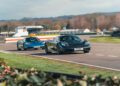Two sleek sports cars, one black and one blue, race on a track near green grass and blurred trees at Goodwood, with "Super Shell" signage visible in the background.