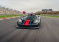 A sleek black sports car, influenced by Gordon Murray's innovative automotive design, zooms down the race track with red and blue stripes. The blurred surroundings reveal hints of buildings in the background, reminiscent of a high-speed dash at Goodwood.