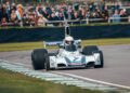 A vintage Formula 1 car with Martini Racing livery speeds around the Goodwood track, showcasing exceptional automotive design, as spectators watch in awe.