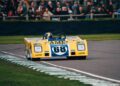 A sleek yellow race car emblazoned with number 68 speeds down the track, the driver expertly handling the wheel beneath his helmet. Spectators at Goodwood cheer from behind the fence, admiring both the thrill of speed and impeccable automotive design.