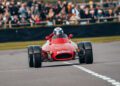 A vintage red race car, reminiscent of Gordon Murray's legendary automotive design, speeds down the Goodwood track, with a helmeted driver at the wheel. In the background, a blurred crowd cheers enthusiastically from behind the barrier.
