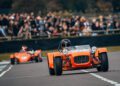 Two orange race cars, reminiscent of Gordon Murray's impeccable automotive design, speed around the track as a sea of spectators watch in awe.