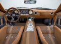 The vintage car interior, reminiscent of a Ford GT, features brown leather seats and dashboard, round gauges, and a left-hand steering wheel. A central gear shift lever and rearview mirror add to its charm.