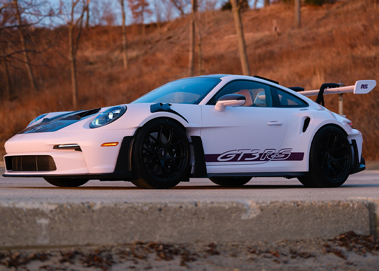 Mobil sport Porsche 911 GT3 RS yang ramping "GT3 Rs" Terpamput di samping, roda hitam, dan sayap belakang besar berdiri di atas permukaan beton, dibingkai oleh siluet pohon telanjang.