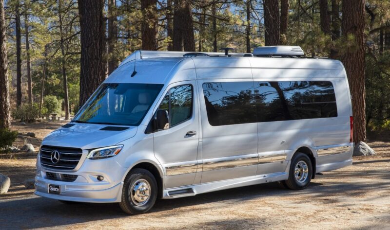 A white Mercedes-Benz Sprinter, a unique creation parked in a wooded area, showcases its left side and front with large windows and an external AC unit on the roof.