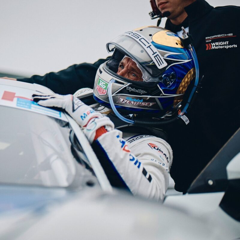 A race car driver in a helmet and suit prepares for a star turn, entering a Porsche, assisted by someone in a Wright Motorsports jacket.