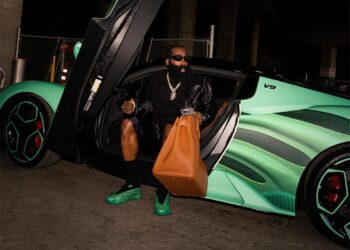 A person sits in a sleek, green Maserati MC20 with its upward door ajar, dressed in a black outfit and sunglasses. Clutching a large brown bag, they exude an air of mystery as if arriving at an exclusive sneaker release event. The intricate wheel designs complement the scene perfectly.