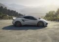 A sleek silver Lotus Emira sports car is parked on a scenic mountain overlook, surrounded by trees and distant hills under a hazy sky.