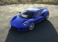 A sleek blue sports car, the Lotus Emira, is parked on a paved area surrounded by a low stone wall, with trees gracefully lining the background.