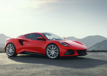 A sleek red sports car, the Lotus Emira Turbo, rests on a paved surface with majestic mountains in the backdrop under a clear sky, boasting an impressive 400HP.