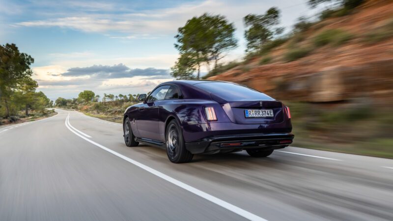 A purple luxury coupe driving on a winding road through a scenic landscape with trees and hills under a partly cloudy sky.