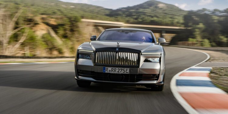 A silver car with license plate "RR 275E" is driving on a curved road with hills and trees in the background.