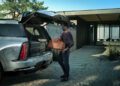 A man wearing sunglasses loads an orange duffel bag into the open trunk of a parked 2026 Escalade in a modern driveway setting, showcasing the sleek design and advanced technology Cadillac's IQL promises.