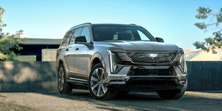A sleek silver 2026 Escalade IQL sits on a gravel surface under a clear blue sky, with trees and a building in the background.