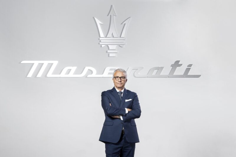 A man in a suit stands confidently with arms crossed in front of a large Maserati logo, echoing the elegance of the Grecale model on a light background.