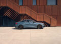 An Audi RS 3 sedan is parked on a wide concrete pavement in front of a modern building with rust-colored walls and a staircase.