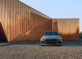 A gray Audi RS 3, renowned for its dynamic 5-cylinder engine, is parked on a cobblestone surface in front of a modern building with copper-colored geometric panels.