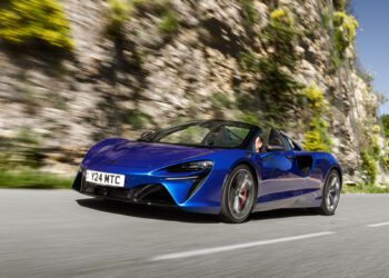 A blue sports car with a convertible top, reminiscent of a McLaren Trophy America series model, drives on a winding road next to rocky terrain and greenery under a clear blue sky.