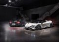 Two sports cars in an industrial setting: a sleek McLaren in white on the right and a dark-colored car on the left. Both are parked on a reflective concrete floor under bright overhead lighting, reminiscent of thrilling LT Cars owner events.