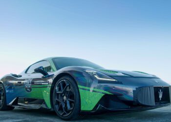 A sleek, dark-colored Maserati MC20 is parked outdoors on a clear day, its visible logos and graphics shining under the sun, hinting at its autonomous driving capabilities.