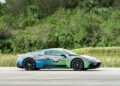 A sleek green and black Maserati sports car, adorned with "NDV" decals, is parked on a road surrounded by lush greenery and towering trees.