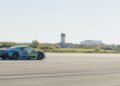 A blue Maserati MC20 with decals races on a wide, empty runway. Amidst a backdrop of industrial buildings and a control tower, it speeds forward on a clear day, pursuing the Autonomous World Speed Record.