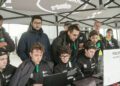A group of people under a tent, focused on laptops. They wear matching black jackets with Maserati logos. A "Politecnico Milano 1863" sign is visible, hinting at their possible pursuit of an Autonomous World Speed Record, perhaps even for the MC20 model.