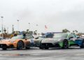 Two stunning Maserati cars, the MC20 in vibrant orange and another in sleek green, stand elegantly on a wet surface adorned with decals and numbers. The scene is set against an overcast sky, with palm trees swaying gently in the background.