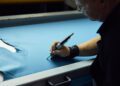 With over 600 hours of dedication, a person wearing a wrist support skillfully uses a tool to draw on blue fabric laid out on the table, reminiscent of the precision and craftsmanship that defines Rolls-Royce.