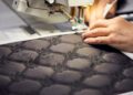 Hands operating a sewing machine, meticulously stitching a dark, intricately-patterned fabric with interlocking designs. Each detail requires hours of build precision, mirroring the craftsmanship found in a Rolls-Royce.