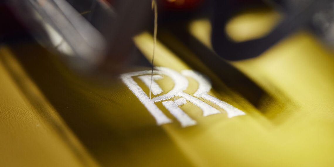 Close-up of a sewing machine embroidering the opulent "RR" initials on yellow fabric, reminiscent of the craftsmanship found in a luxury Rolls-Royce.