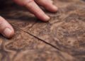 A hand with lightly worn nails inspects two adjoining pieces of richly grained, brown wood veneer featuring intricate swirling patterns, reminiscent of the craftsmanship found in a Rolls-Royce, where build time exceeds 600 hours.