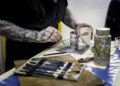 A person with tattooed arms delicately paints on a small canvas, surrounded by art supplies like jars and a palette. Their attention to detail rivals the build time of a Rolls-Royce, where every brush stroke mirrors the precision of crafting a luxury car.