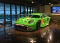 A vibrant green race car with a smiling face design gleams in the showroom at the Porsche Experience Center, Atlanta. This ultra-exclusive lineup boasts a backdrop of sleek screens and expansive windows, enhancing its allure.