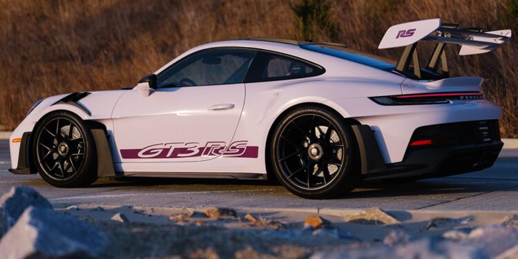 A sleek white 911 GT3 RS with a PTS Moongem finish flaunts its "GT3 RS" decal and large rear wing, resting on black wheels. It's parked stylishly on a dirt road, framed by dry grass, as if fresh from the Porsche Exchange showroom.