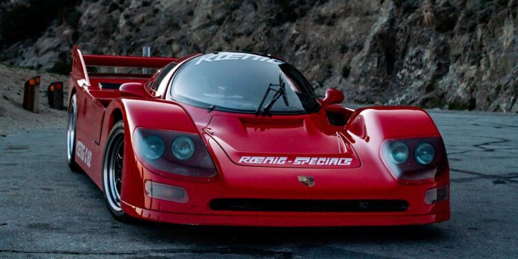 A red sports car with the "Koenig-Special" label proudly displayed on its hood, the C-62 Special is parked on a winding mountain road. Rugged rocky cliffs stand tall in the background, creating a scene fit for a Petrolicious feature.