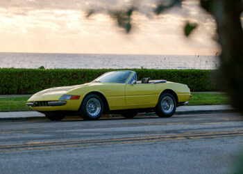 A yellow convertible sits parked by the roadside, embodying a scene straight out of a Daytona Vice episode. With a hedge and the ocean as its backdrop under a partly cloudy sky, it feels like a frame from Petrolicious—a story waiting to be told on New Film Friday.
