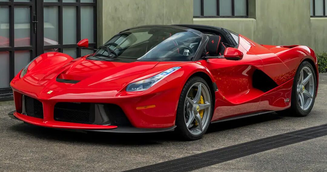 A red Ferrari sports car with a convertible roof, one of the most expensive Ferraris, is parked on a paved surface in front of a modern building with large windows.