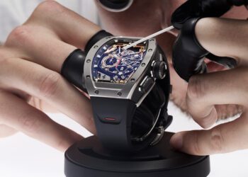 A person wearing black gloves is meticulously inspecting a Richard Mille luxury watch with a blue face and visible gears, carefully using a tool while it's mounted on a stand.