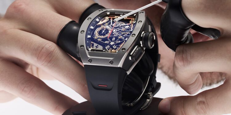A person wearing black gloves is meticulously inspecting a Richard Mille luxury watch with a blue face and visible gears, carefully using a tool while it's mounted on a stand.