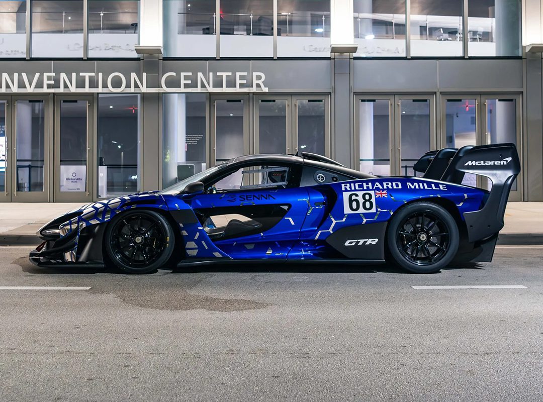 A blue McLaren Senna GTR, inspired by Ayrton, with the number 68, is parked on a city street in front of a convention center at night.