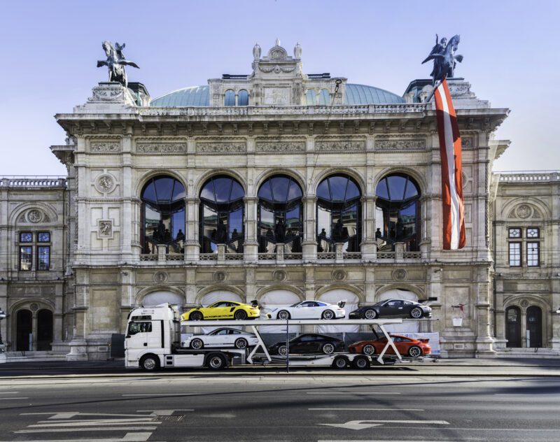 A car transport truck loaded with the exclusive Porsche Collection is parked in front of an ornate, historical building adorned with arches and statues, ready for delivery.