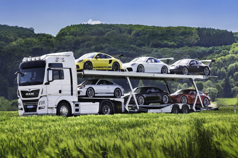 A car transporter truck hauls a stunning Porsche collection of six sports cars through the scenic countryside, with green fields and rolling hills providing a picturesque backdrop.