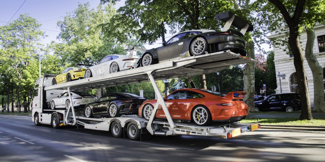 A car transporter truck is carrying six luxury sports cars, including the exclusive Porsche Collection, three on the upper deck and three on the lower, as it cruises down a tree-lined urban street.