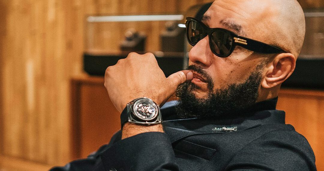 Bald man with sunglasses sits in a chair, hand on chin, exuding Swizz Beatz's style. He's wearing a black suit and a striking De Bethune GMT watch. Behind him, a wooden wall and blurred objects complete the scene.