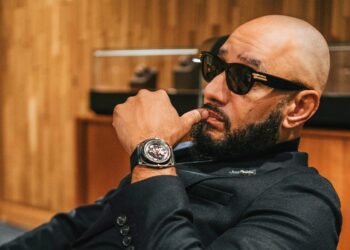 Bald man with sunglasses sits in a chair, hand on chin, exuding Swizz Beatz's style. He's wearing a black suit and a striking De Bethune GMT watch. Behind him, a wooden wall and blurred objects complete the scene.