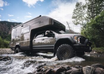 An adventure-ready expedition vehicle drives through a shallow rocky stream, surrounded by trees and mountains under a cloudy sky.